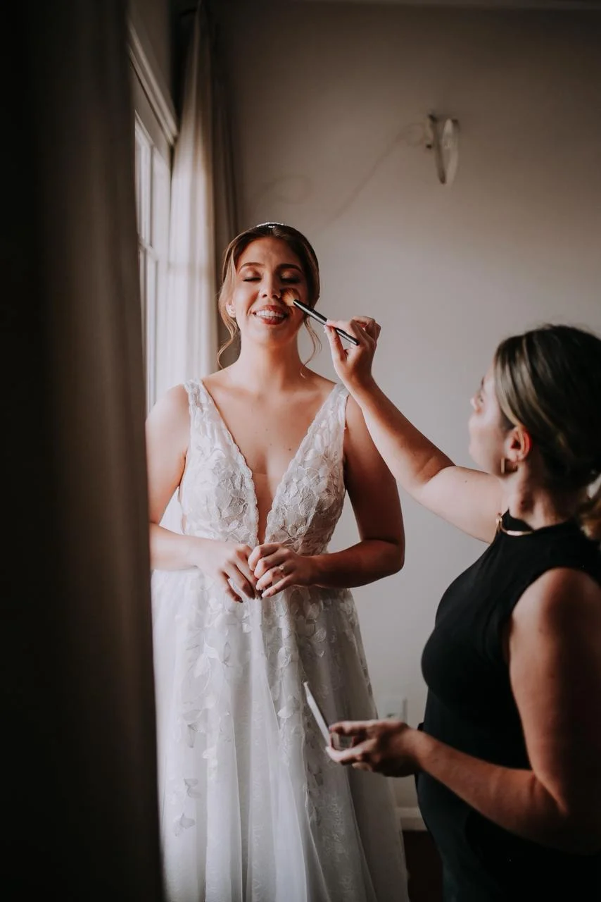 Karol Carminatti maquiando noiva junto à janela com luz natural no dia do casamento